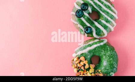 Ciambelle vegane con semi di chia condite con glassa di tè matcha sana con pistacchio e mirtillo. Ciambelle da tè matcha verdi su sfondo rosa. Spazio di copia. Vista dall'alto. Banner Foto Stock