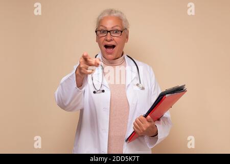 Donna ispanica anziana che indossa uniforme medico tenendo carte e puntando a te con l'indice Foto Stock