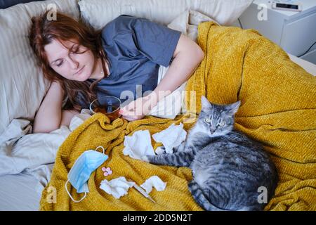 Donna rossa con virus influenzale beve tè sul letto accanto a un gatto Foto Stock