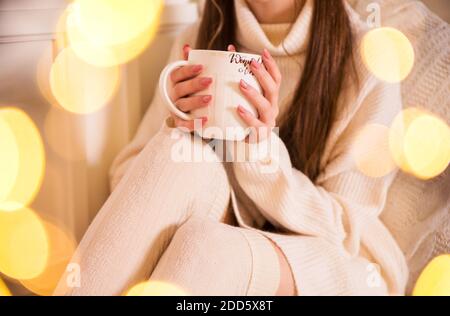 Una ragazza in un maglione bianco e leggings tiene una tazza bianca grande nelle sue mani Foto Stock