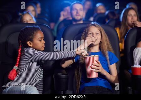 I bambini che si divertono insieme, il pubblico multirazziale giovane sullo sfondo. Amici felici seduti al cinema, guardando fumetti divertenti, caucasica ragazza mangiare e condividere pop corn con l'amica africana. Foto Stock