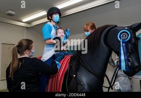 East Lothian, Scozia, UK, NHS Lothian & Muirfield Riding Therapy festeggia 10 anni di ippoterapia: Il fisioterapista Heather Falconer ha aperto la strada al Children’s Therapeutic Riding Service, che fornisce gratuitamente un'equitazione terapeutica a bambini, giovani e adulti con disabilità. Il servizio è stato il primo in Scozia per i bambini con problemi di mobilità, equilibrio, tono muscolare e coordinazione. Oreo, un cavallo meccanico, permette ai giovani di divertirsi con la fisioterapia. Jamie Burns, di 2 anni, cavalca il simulatore meccanico di cavalli Racewood con Lizzie, fisioterapista Foto Stock