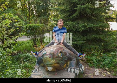 un allegro ragazzo sorridente in una t-shirt a righe si siede su un statua di dinosauro nel parco giochi Foto Stock