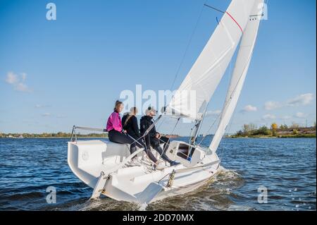 Un bellissimo yacht bianco da corsa monomarca sta navigando contro un bellissimo paesaggio fluviale con un cielo blu. Un uomo e due ragazze sono a bordo Foto Stock