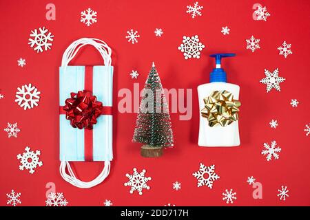 Decorazioni natalizie con maschere facciali monouso e disinfettante per le mani su sfondo rosso. Natale e Coronavirus Pandemic concetto. COVID-19. Foto Stock