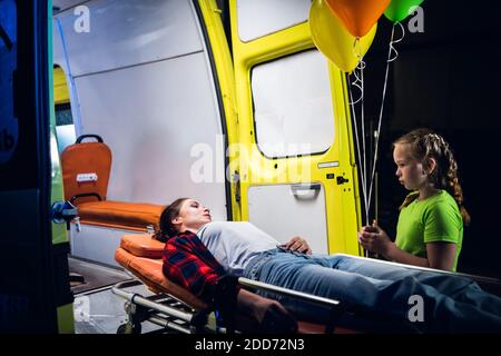 Bambina in piedi accanto a una donna che si trova su una barella accanto a un'ambulanza, si guardano l'un l'altro. Foto Stock