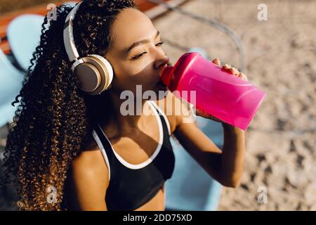 Giovane atletica afroamericana indossando cuffie che riposano dopo intenso fare jogging e bere acqua da una bottiglia durante una pausa dopo jogging o Foto Stock