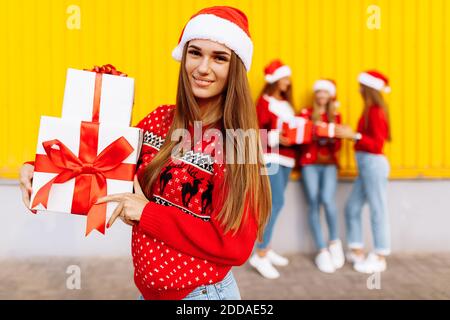 Bella giovane donna sorridente che indossa il cappello di babbo natale con natale presente in piedi sullo sfondo di amici felici che celebrano il natale Foto Stock