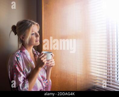 Donna giovane premurosa in accappatoio guardando attraverso la finestra mentre ha caffè a casa Foto Stock