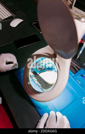 Uomo esperto da circuito stampato di telefono cellulare visto attraverso lente d'ingrandimento presso il centro di riparazione Foto Stock