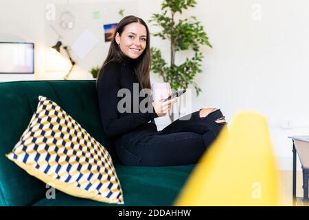 Blogger femmina sorridente che tiene smartphone e tazza di caffè mentre posto a sedere su un divano nell'appartamento loft Foto Stock