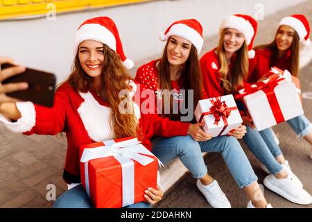 Gruppo di giovani donne in babbo natale cappelli in regalo scatole e presa selfie sul telefono cellulare mentre si siede sopra marciapiede all'aperto Foto Stock