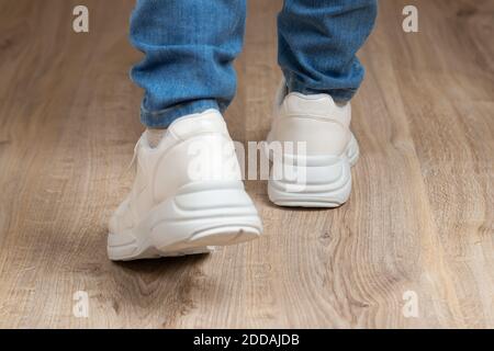 Vista posteriore dei piedi calpestati in sneaker bianche con suola a pezzi e jeans blu sul pavimento marrone. Un paio di nuove scarpe comode per uno stile di vita attivo. Foto Stock