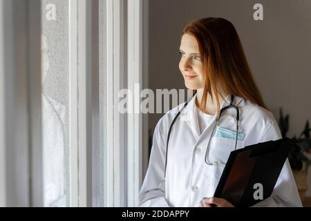 Premuroso giovane dottore sorridente guardando attraverso la finestra dentro ufficio domestico Foto Stock