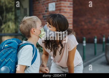 Madre che baciano il figlio indossando una maschera protettiva in piedi in font di scuola Foto Stock