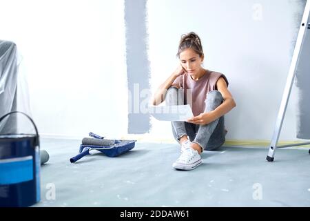 Giovane donna con testa in mano che tiene carta mentre si siede sul pavimento a casa Foto Stock