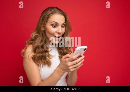 Immagine di una bella donna bionda scioccata giovane in posa isolata su sfondo rosso muro utilizzando il telefono cellulare. Foto Stock