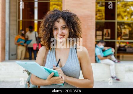 Giovane donna sorridente con documenti in piedi nel campus universitario Foto Stock