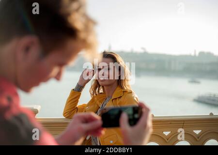Giovane uomo che fotografa una giovane donna con uno smartphone sul ponte contro il cielo Foto Stock