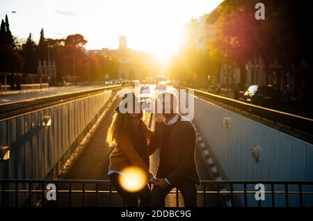 Coppia che tiene le mani mentre si siede sulla ringhiera del ponte insieme a. città durante il tramonto Foto Stock