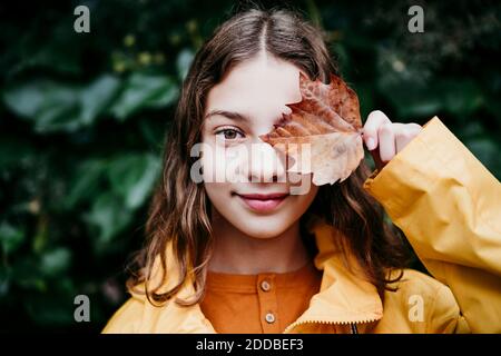 Sorridente ragazza in impermeabile occhione con foglia di edera secca mentre si sta contro il muro di foglia Foto Stock