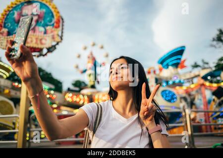 Bella giovane donna che prende selfie attraverso smartphone mentre gesturing cartello di pace al parco divertimenti durante l'estate Foto Stock