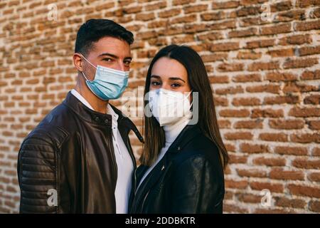 Giovane coppia che indossa maschera faccia in piedi insieme contro muro di mattoni Foto Stock