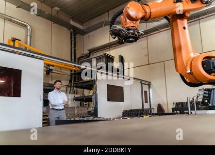 Uomo d'affari con braccia incrociate che monitorano il braccio automatico del robot mentre è in piedi in fabbrica Foto Stock