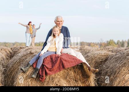 Nonno e nipote seduti sulla balla di fieno mentre nonna e. nipote in background Foto Stock