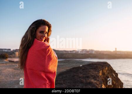 Donna sorridente coperta di scialle in piedi in spiaggia Foto Stock