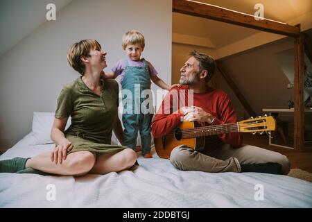 Figlio in piedi tra madre e padre che suona la chitarra seduto a casa in camera da letto Foto Stock