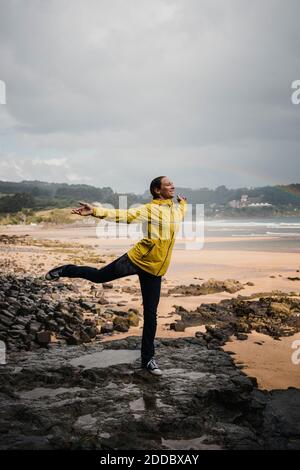 Giocosa donna in impermeabile in piedi su una gamba a. spiaggia durante la stagione delle piogge Foto Stock