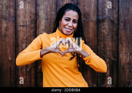 Donna matura sorridente che forma il cuore con la mano mentre si è in piedi contro muro di legno Foto Stock