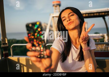 Bella giovane donna che prende selfie attraverso il telefono cellulare mentre ridendo E segno di pace gesturante sulla ruota di Ferris Foto Stock