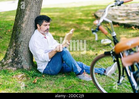 Libro di lettura dell'uomo maturo con concentrazione mentre sedendo in pubblico parcheggio Foto Stock