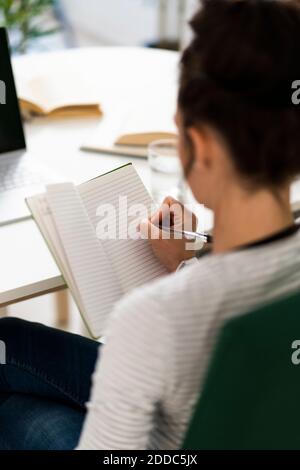 Donna mid adulta che scrive in blocco note mentre si siede sulla sedia a casa Foto Stock