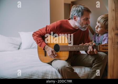 Padre che suona la chitarra davanti al figlio mentre si siede letto a casa Foto Stock