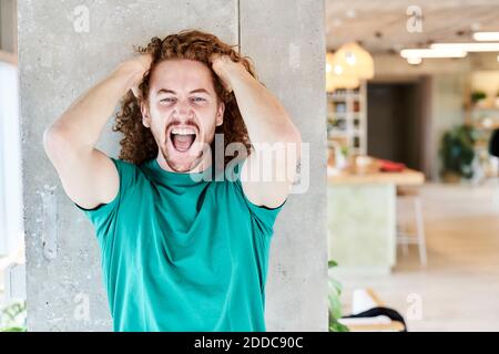 Uomo arrabbiato con le mani in capelli gridando mentre in piedi contro colonna in soggiorno a casa Foto Stock