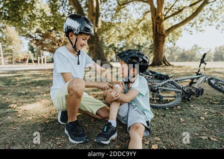 Ragazzo sorridendo dopo aver messo il bendaggio sul ginocchio del fratello più giovane seduto nel parco pubblico Foto Stock