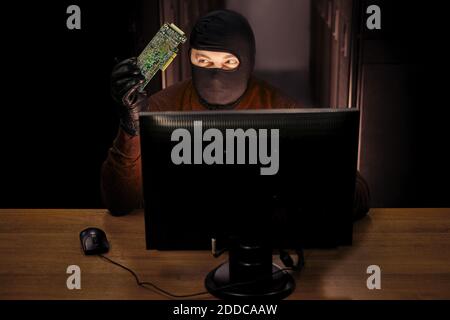 Uomo in maschera nera con una scheda a circuito stampato da un dispositivo hacker in mano si siede a un computer. Concetto di problemi di sicurezza dei dati e di hacking della rete Foto Stock