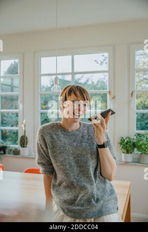 Donna sorridente che invia messaggi vocali tramite smartphone mentre si è in piedi casa Foto Stock