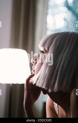 Giovane donna con la mano sul mento in piedi da una lampada illuminata a casa Foto Stock