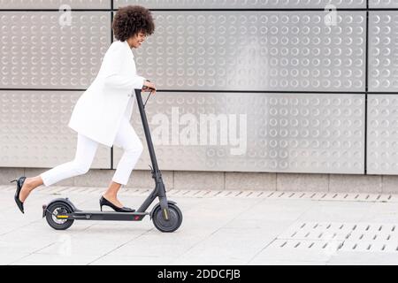 Donna d'affari sorridente che guida scooter elettrico a spinta contro parete argentata Foto Stock