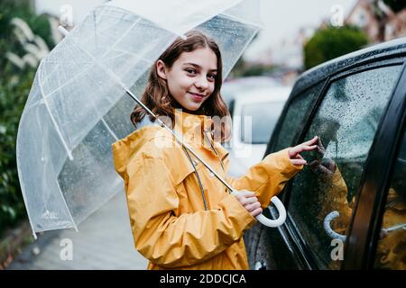 Sorridente ragazza in impermeabile che tiene l'ombrello mentre si è in piedi in auto in città Foto Stock