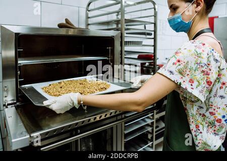 Giovane donna panettiere posizionamento foglio in forno a panetteria cucina durante la pandemia Foto Stock