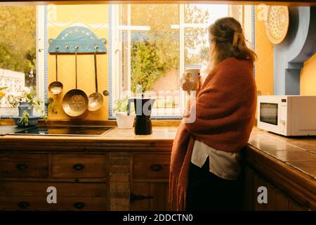 Donna anziana in coperta che guarda attraverso la finestra mentre beve caffè a casa Foto Stock