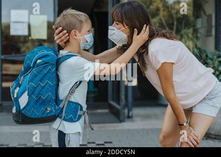 Madre e figlio indossano una maschera protettiva in piedi faccia a faccia di fronte all'edificio scolastico Foto Stock