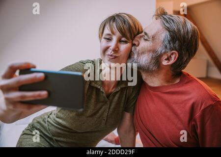 Uomo maturo che bacia la donna mentre prende selfie sullo smartphone seduto in camera da letto a casa Foto Stock