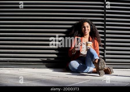 Donna sorridente che tiene una tazza riutilizzabile e uno smartphone mentre si è seduti contro l'otturatore nero Foto Stock