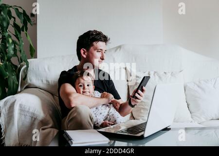 Messaggi di testo Padre su smartphone che tiene il figlio mentre si siede sul divano a casa Foto Stock